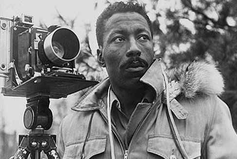 Portrait of Gordon Parks standing next to a camera on a tripod